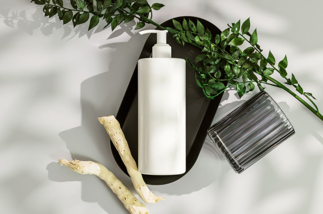 Top view of a white cosmetic bottle with pump surrounded by green leaves and wood on a white surface.